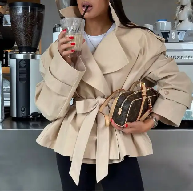 Жінка у бежевому весняному пальті-тренчі з поясом тримає сумку та напій у кав’ярні